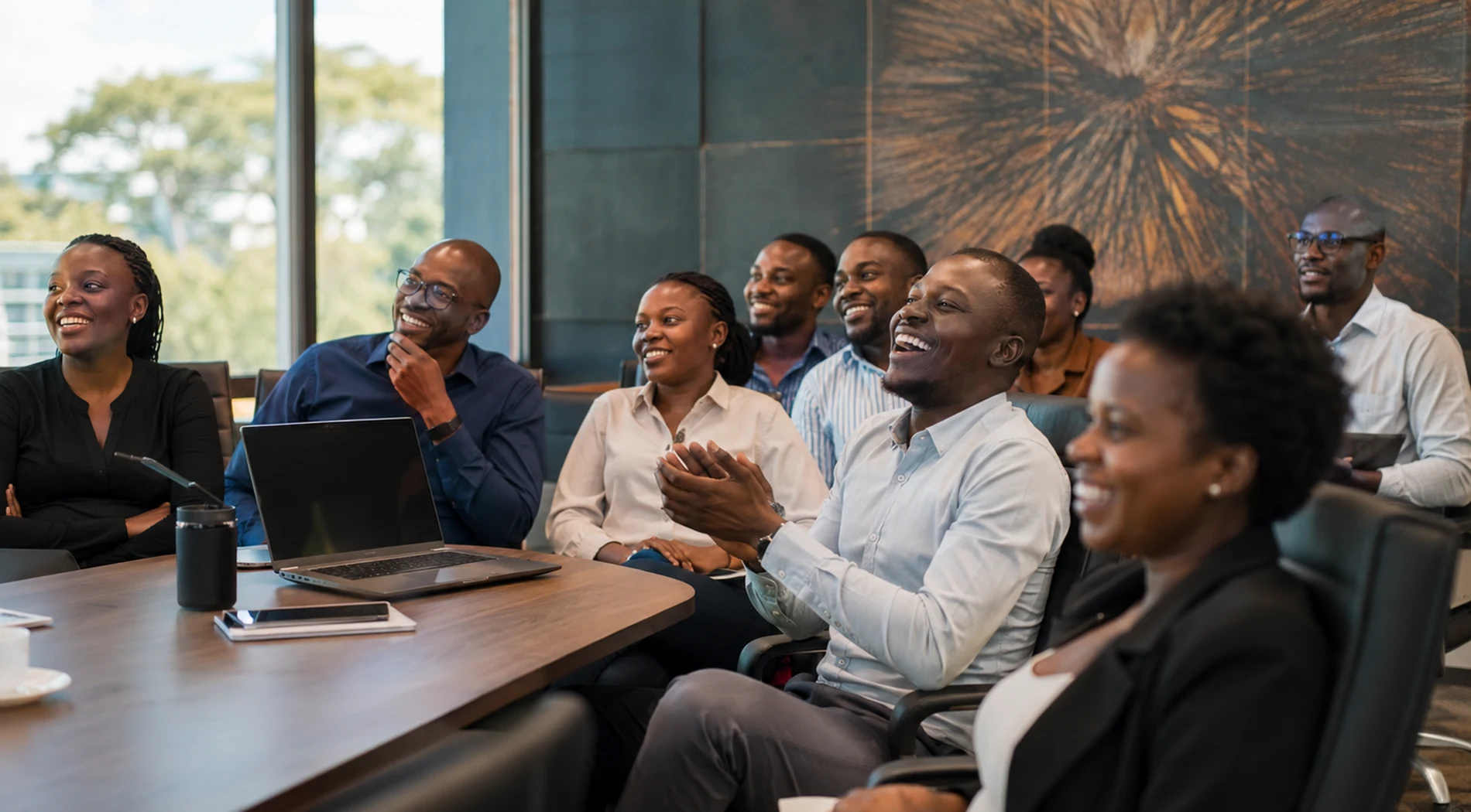 A Zimbabwean office team gathered in a meeting room, laughing and applauding at a video on the screen