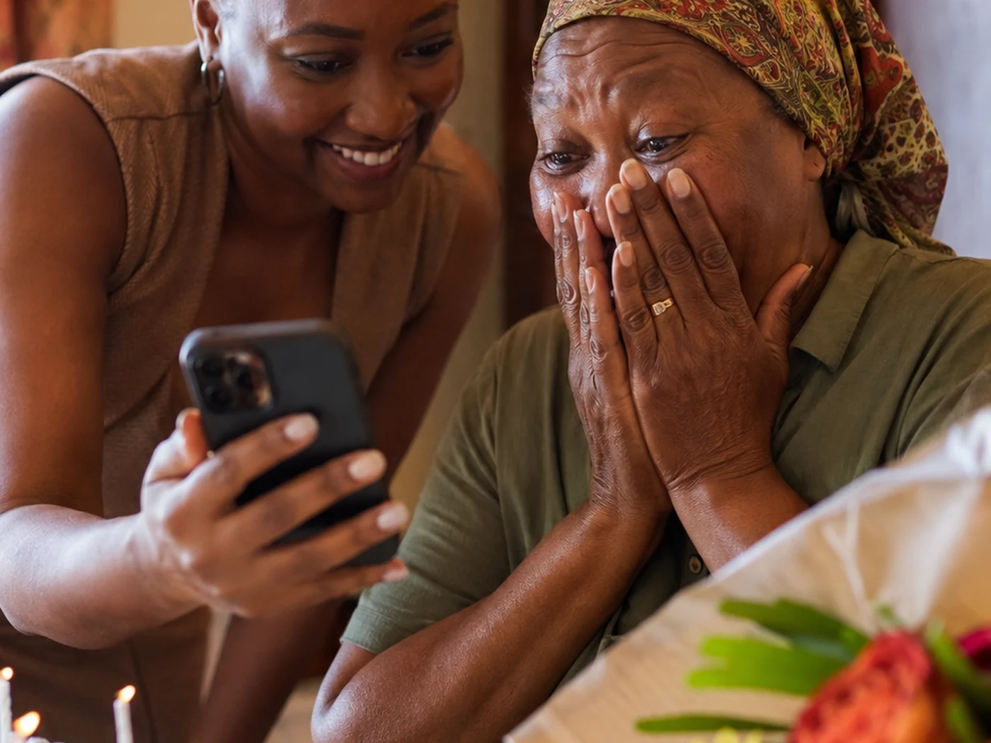 An older Zimbabwean woman, hands to her cheeks, watching a personal video message her daughter is holding up